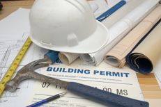 A building permit on a table, with building plans, tools and a hard hat sitting on top
